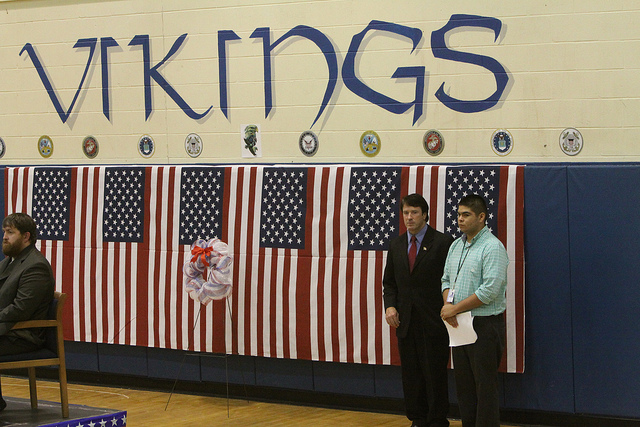 A student and a man standing in the gym with US Flags hanging behind them and VIKINGS painted on the top